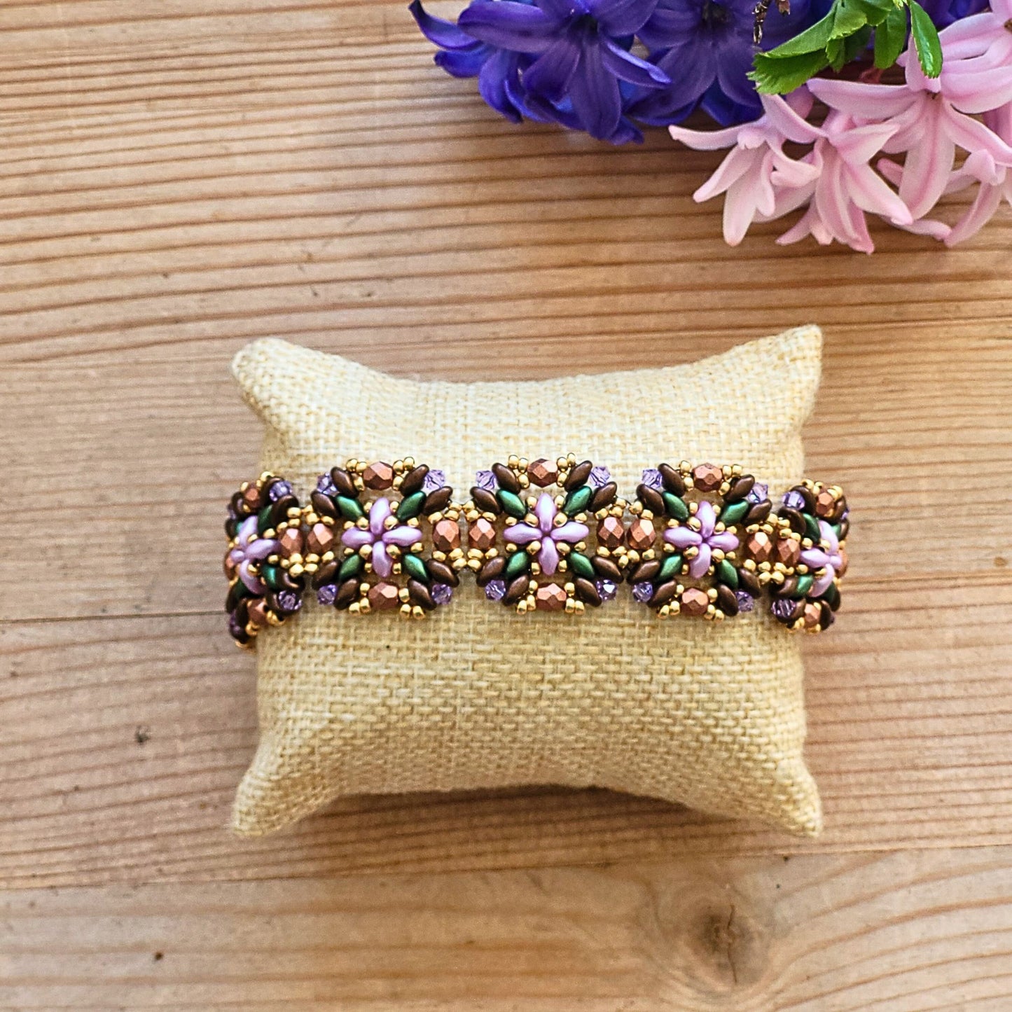 Håndlavet armbånd med blomsterformet mønster i perler – en farverig kombination af grøn, lilla, rosa og gylden på naturfarvet pude, med forårsblomster i baggrunden