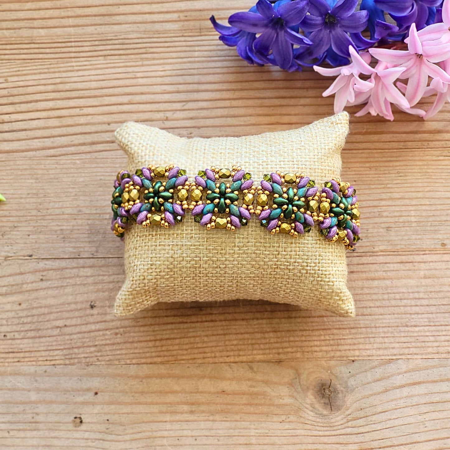 Håndlavet armbånd med blomsterformet mønster i perler – en farverig kombination af grøn, lilla, rosa og gylden på naturfarvet pude, med forårsblomster i baggrunden