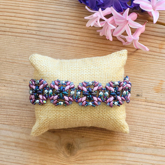 Håndlavet armbånd med blomsterformet mønster i perler – en farverig kombination af lilla, lilla, solv og rosa på naturfarvet pude, med forårsblomster i baggrunden