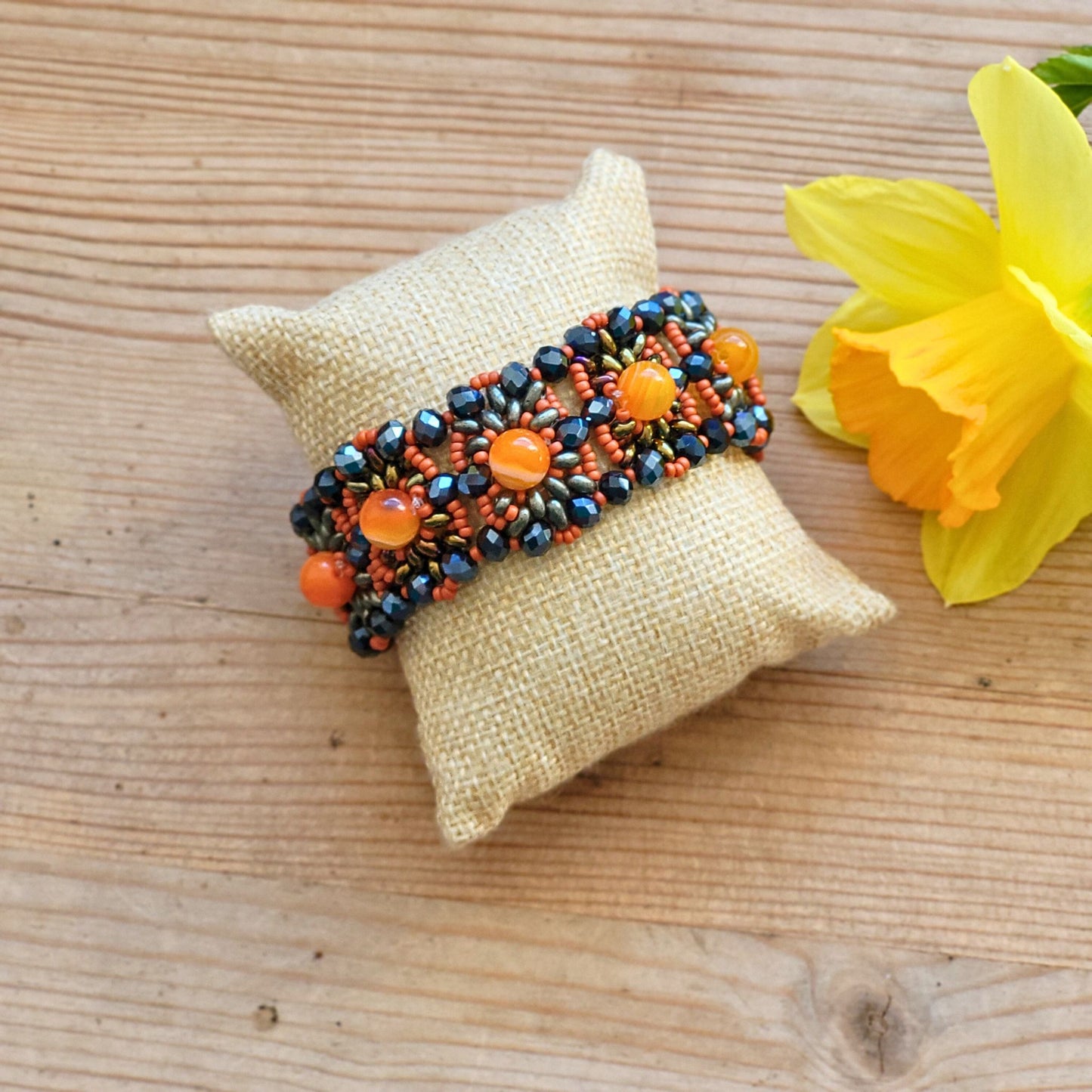 Håndlavet armbånd i Vintage-stil med naturlige halvædelsten i varme orange og koralfarver, kombineret med glasperler i blå og bronze, lagt på en lys pude med en påskelilje i baggrunden
