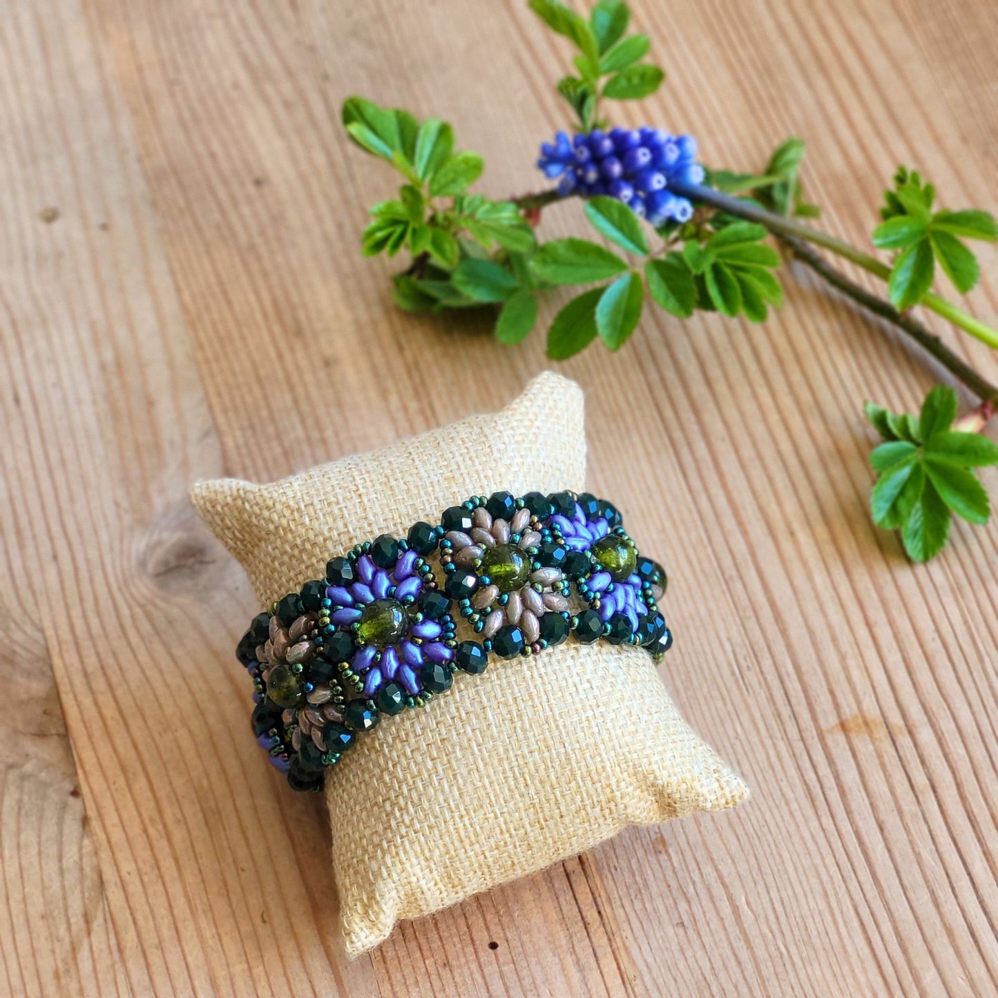 Håndlavet armbånd i Vintage-stil med blomsterformede glasperler i grøn, lilla og beige, dekoreret med små perler i metallic blå, præsenteret på en lys pude med en forårsgren i baggrunden.