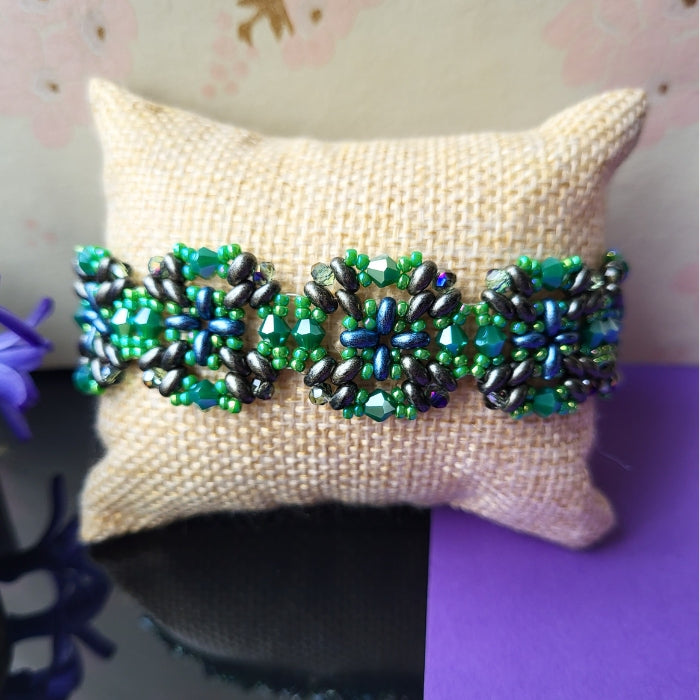 Håndlavet armbånd med blomsterformet mønster i perler – en farverig kombination af grøn, blå og grå på naturfarvet pude, med forårsblomster i baggrunden
