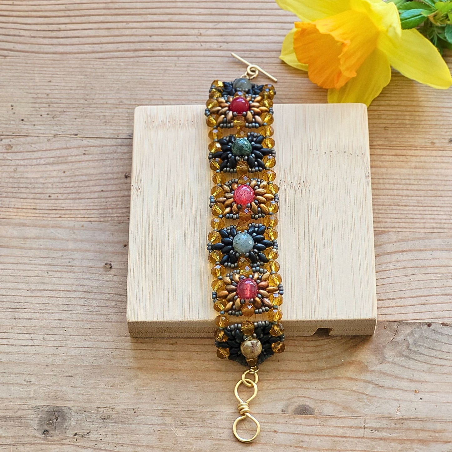 Vintage armbånd håndlavet med glasperler i gyldne og mørke toner og farverige natursten i rød, grøn og blå, præsenteret på en lys træplade med en gul forårsblomst i baggrunden.