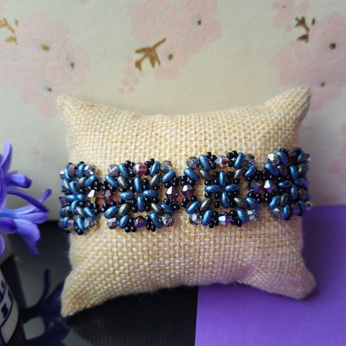 Håndlavet armbånd med blomsterformet mønster i perler – en farverig kombination af b, lilla, grå og sort på naturfarvet pude, med forårsblomster i baggrundenå