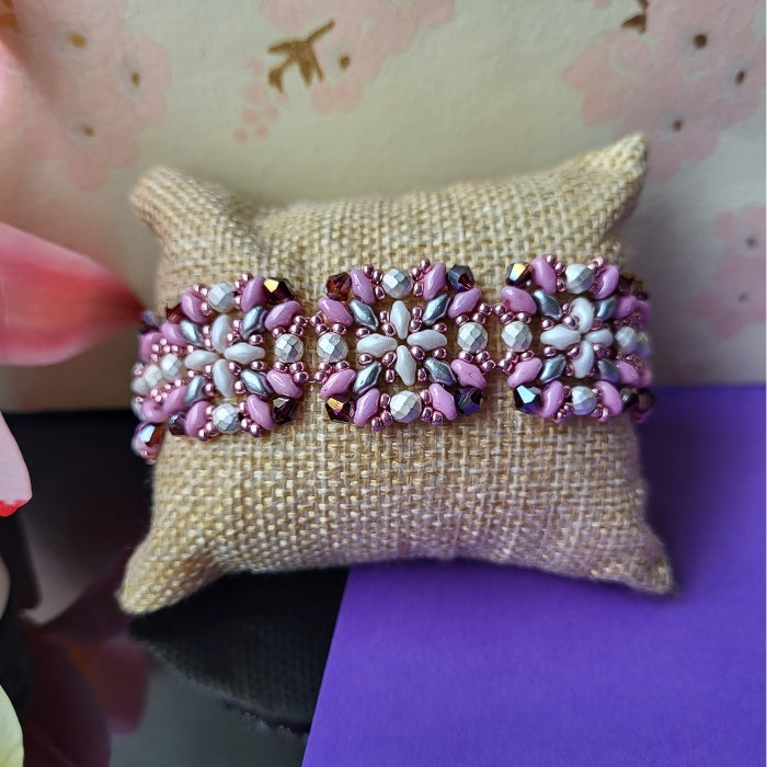 Håndlavet armbånd med blomsterformet mønster i perler – en farverig kombination af sølv, lilla, og rosa på naturfarvet pude, med forårsblomster i baggrunden